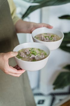 Close-up of hands holding two bowls of delicious homemade soup garnished with herbs.