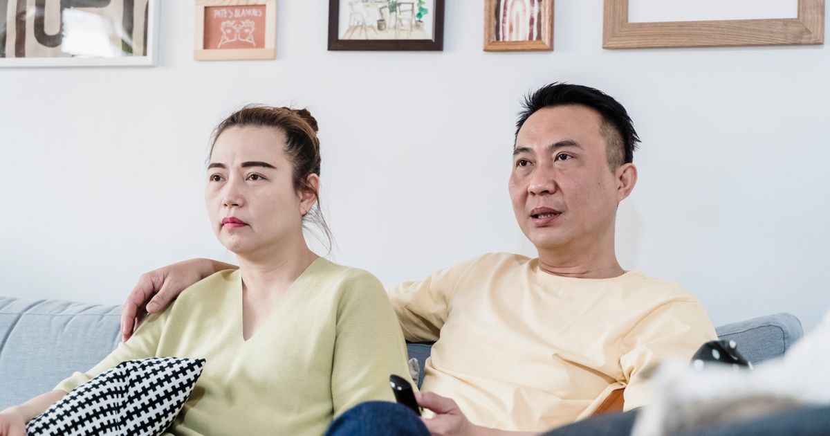 A couple sitting on a couch watching television, one holding a remote.