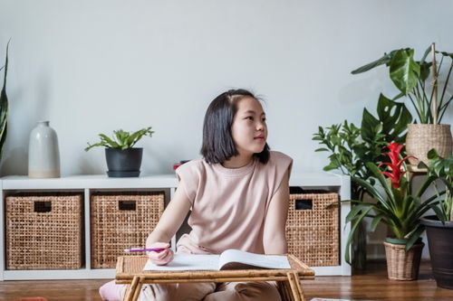 Free Asian girl focused on homework at a small table, surrounded by indoor plants in a cozy room. Stock Photo