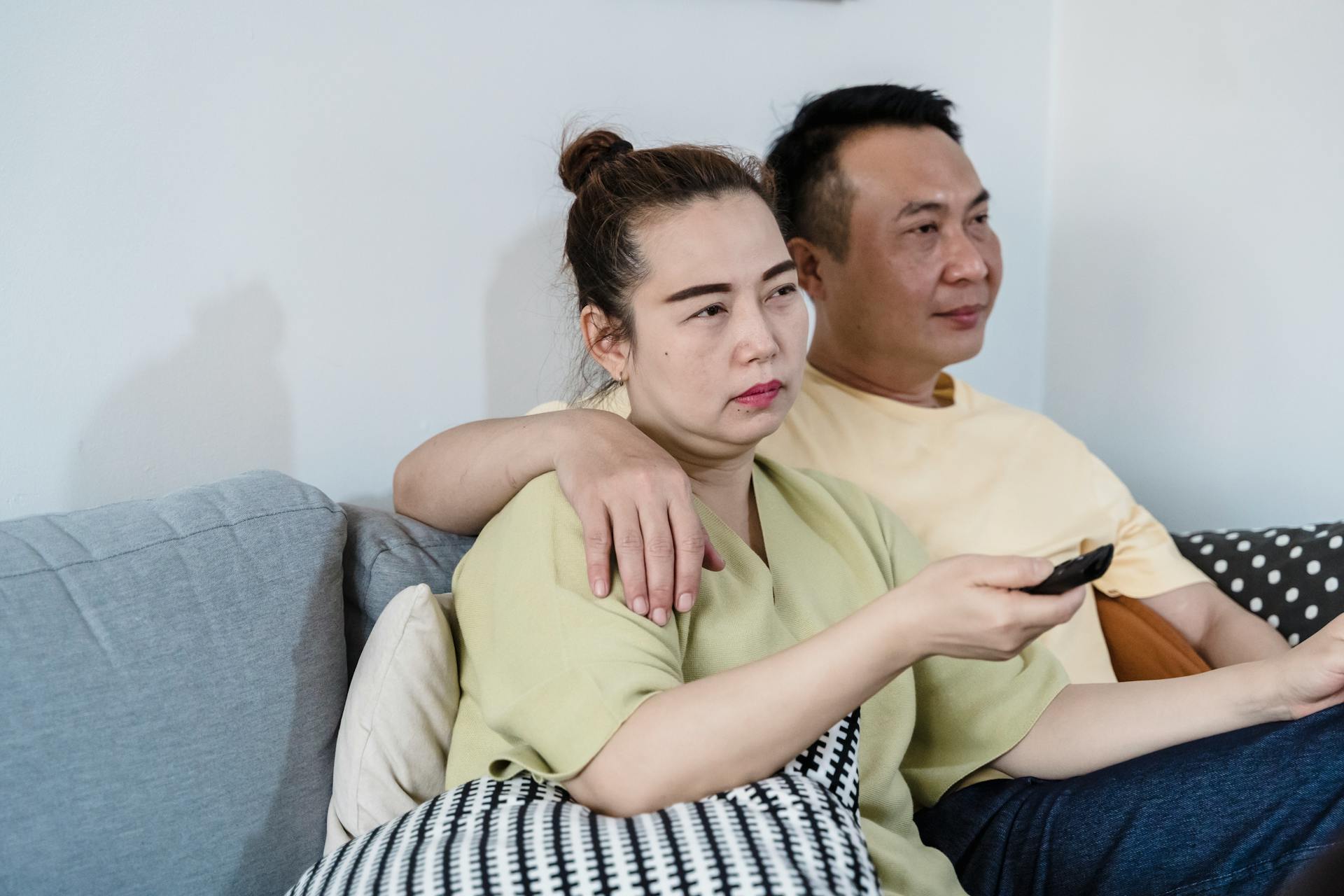 An Asian couple sitting on a couch at home, watching TV together with a remote control in hand.