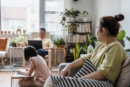 A woman observes while family members engage in work and drawing activities in a sunny living room.