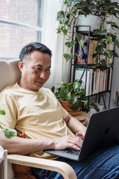 Adult man comfortably seated with a laptop in a cozy, plant-filled living room.