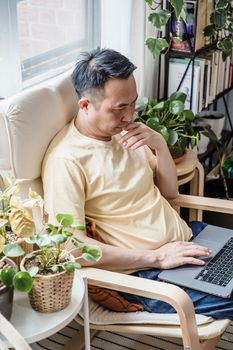 Mature man engaged in remote work, surrounded by lush indoor plants by a sunny window.