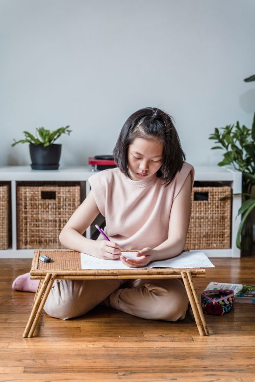 Free Teenager studying at home on a wooden floor, focused on her notes with a small table. Stock Photo