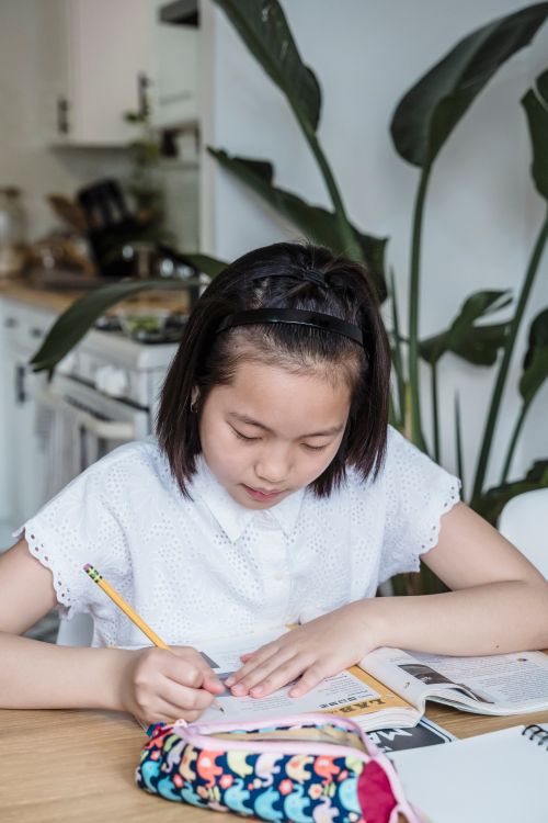 Free A young girl writing on paper while studying at a wooden table in a modern kitchen setting. Stock Photo