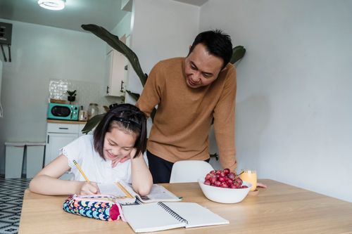 Free Asian father helping his daughter with homework at the kitchen table, fostering education and smiles. Stock Photo