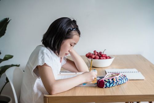 Free Child focuses on homework at a wooden table indoors, surrounded by study materials. Stock Photo