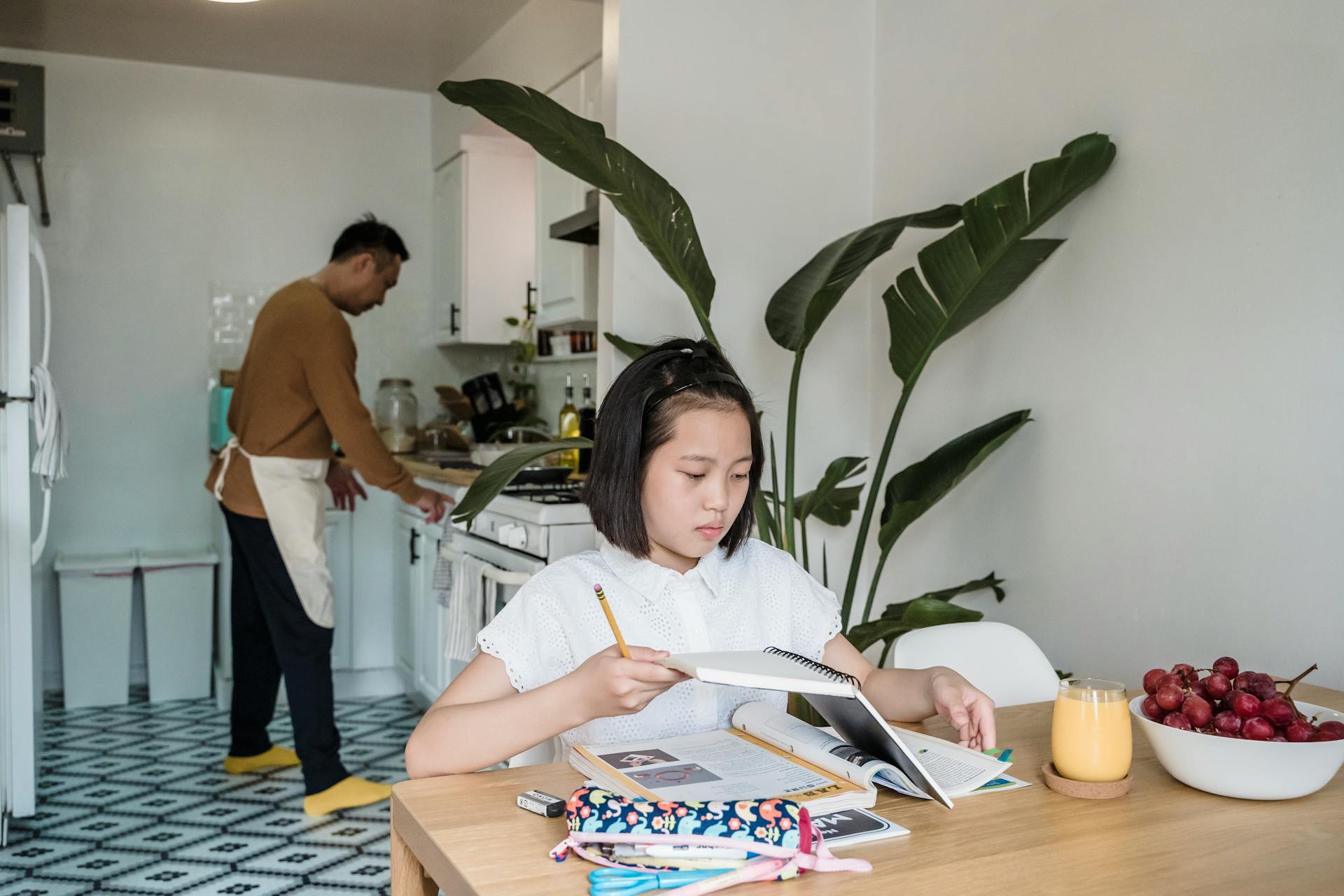 A child focuses on homework at the dining table with a parent cooking in the kitchen.