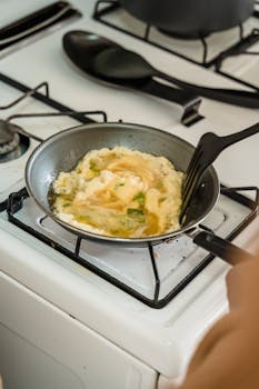 Close-up of scrambled eggs cooking in a frying pan on a gas stove with a spatula.