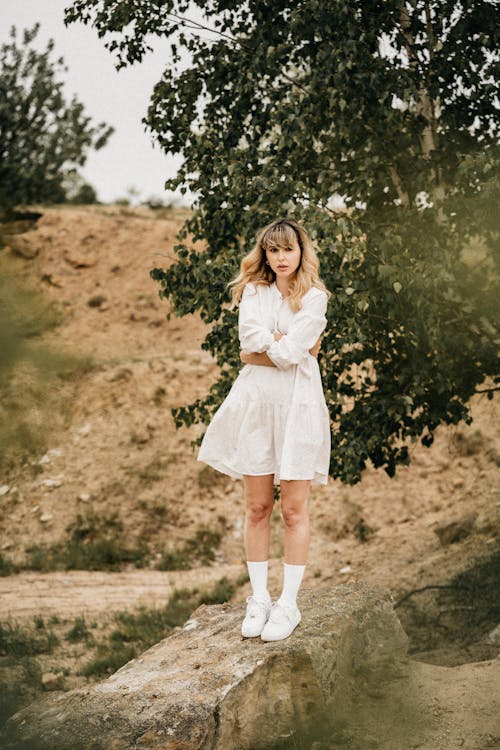 Full body of pensive lady with blond hair in white dress standing with crossed arms on stone near green tree growing on sandy slope of hill in nature in daytime