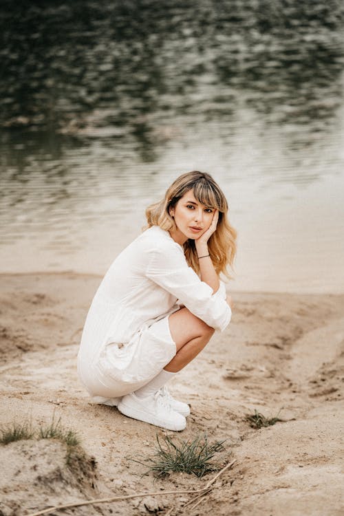 Side view of calm female with blond hair in white dress squatting down and leaning on hand on sandy coast against clam water of lake in nature in daytime