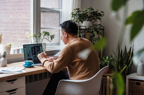 Free A man working from home surrounded by plants, using a laptop at a wooden desk. Stock Photo