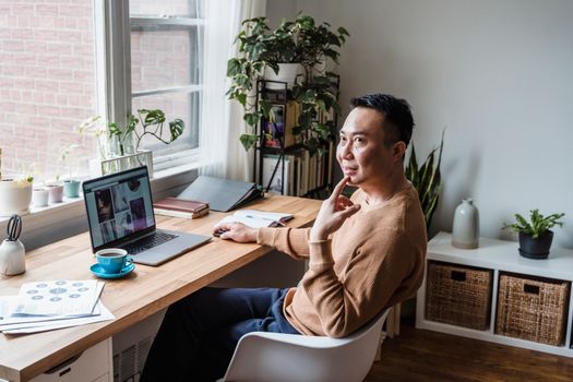 Asian man sitting at a desk with a laptop, coffee cup, and plants, working remotely indoors.