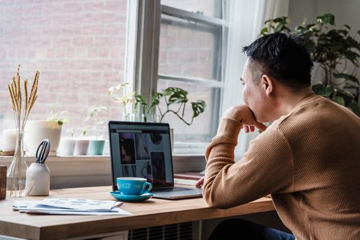 A man in a brown sweater works from home at a desk with a laptop near a window.
