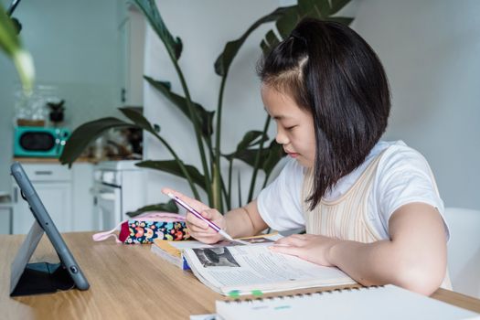 Young Asian girl studying indoors, using a tablet, surrounded by plants and a colorful pen case.
