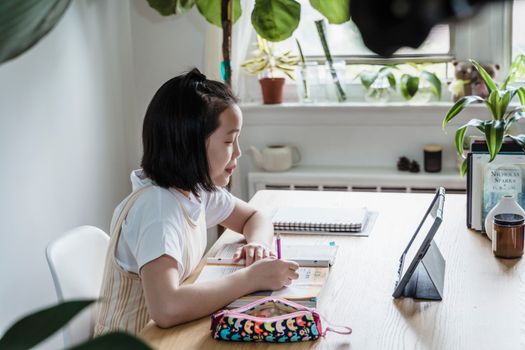 Focused young girl studying online at home with tablet and books on a wooden table.