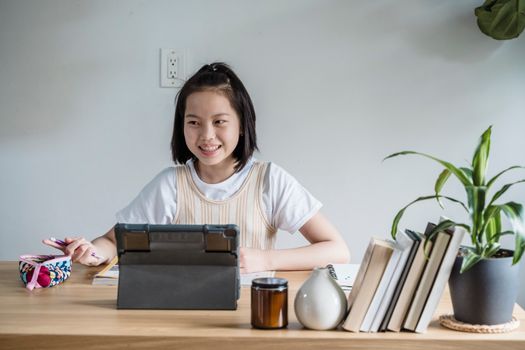 Asian teenager engaged in an online class, using a tablet at a cozy indoor setup.