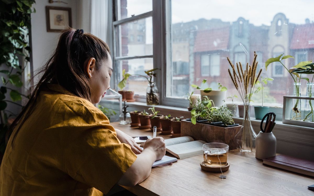 Asian woman writing in notebook in a cozy home office by window with plants.