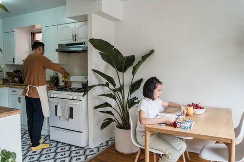 Free Father cooking while daughter studies in a cozy kitchen setting. Stock Photo