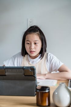 Asian girl focusing on online class using a tablet in a home setting, engaged in learning.