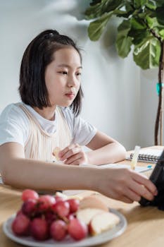 A young girl studies online at home, enjoying healthy snacks of grapes and apples, creating a focused atmosphere.