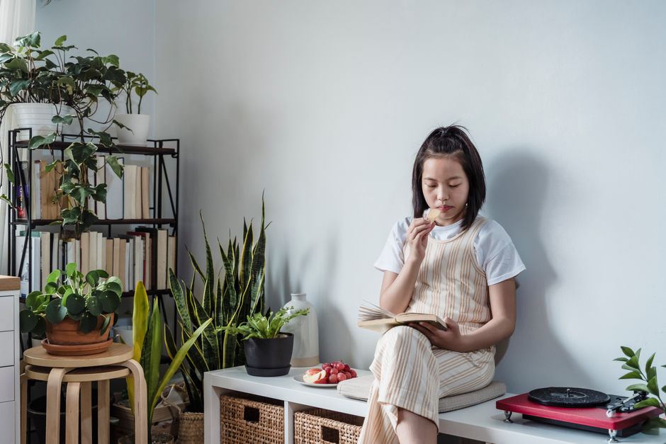 A teenager enjoys a snack while casually reading in a home setting surrounded by indoor plants.