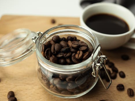 A close-up shot of roasted coffee beans in a glass jar next to a cup of coffee.
