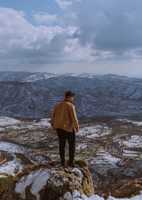 Free A man stands atop a snowy rock, gazing over a vast mountainous landscape under a cloudy sky. Stock Photo