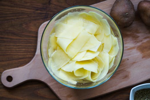 Overhead view of thinly peeled potato slices in a clear bowl on a wooden board.
