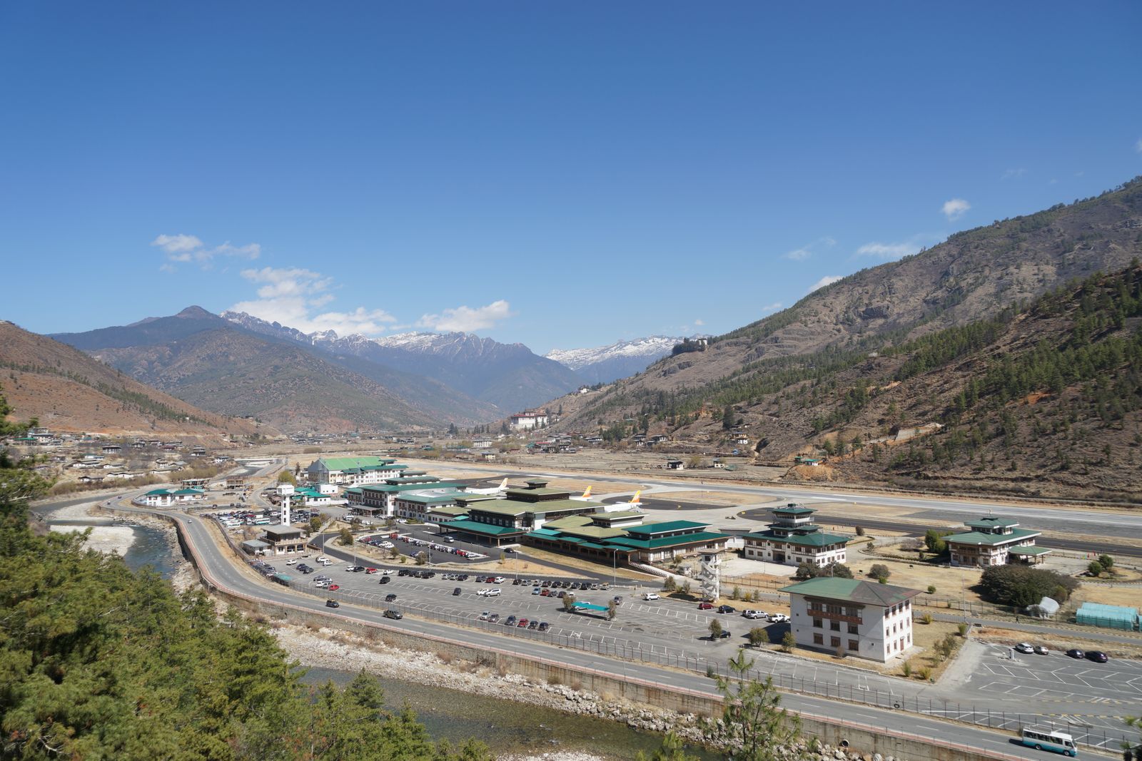 Paro airport's runway and terminal seen from the hillside above, the river alongside and snow peaks at the valley head
