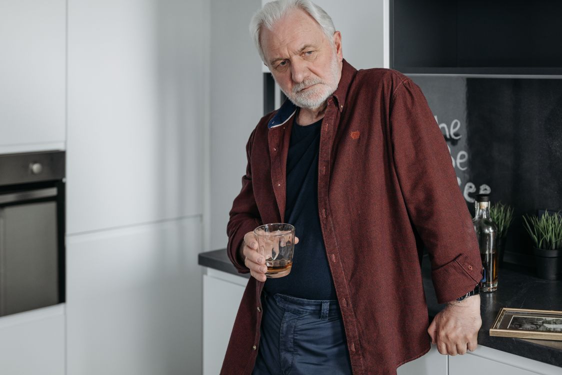Ein älterer Mann in einer Küche, ein Glas in der Hand, in Gedanken versunken.