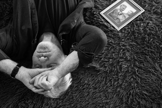 Black and white image of an elderly man lying down, expressing sorrow with a framed photo beside him.