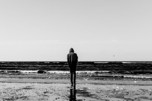 Free A solitary figure in a hoodie stands at the beach, reflecting by the ocean in black and white. Stock Photo