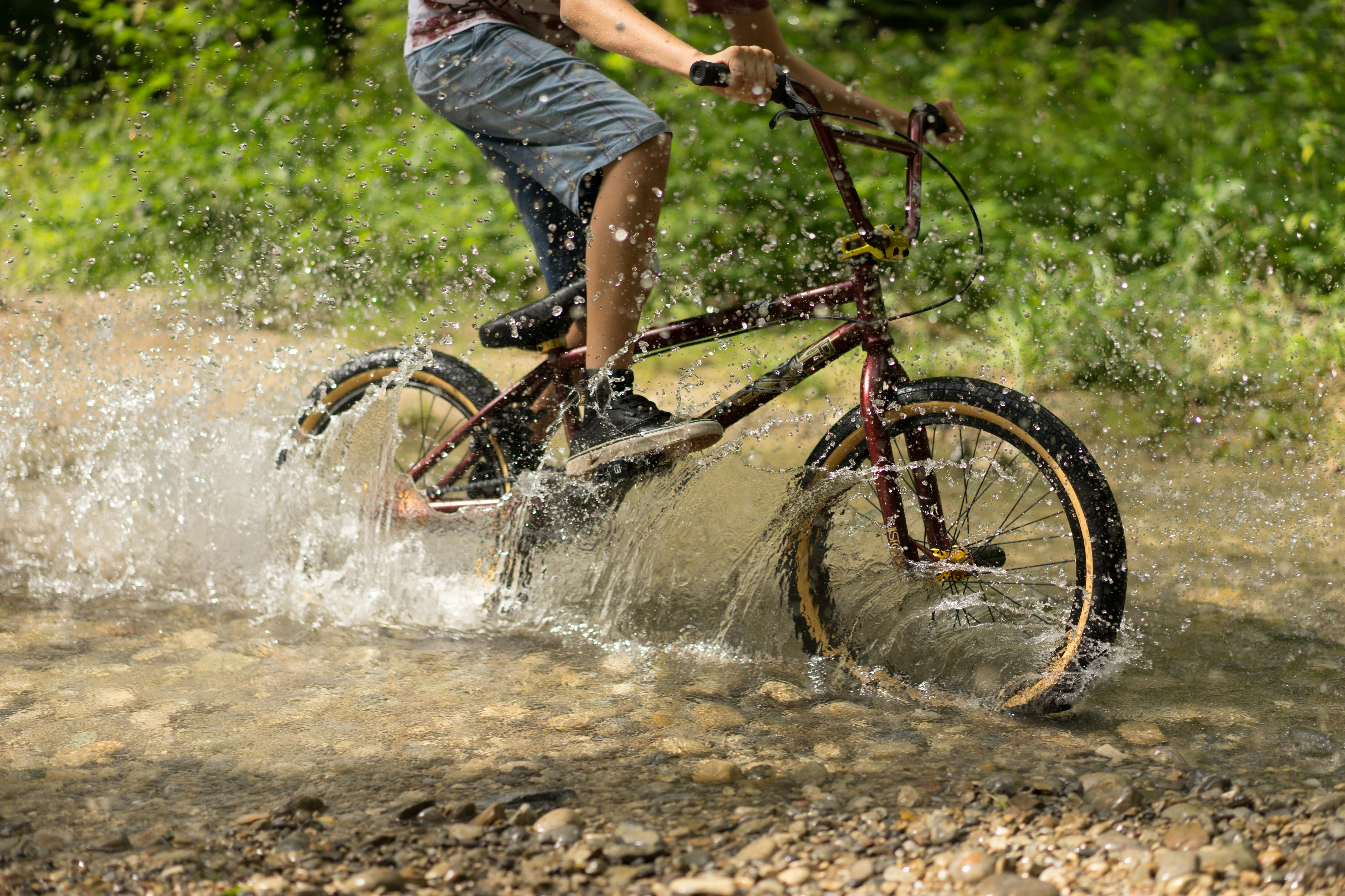 A BMX cyclist rides through a stream in a lush forest, creating a dynamic splash.