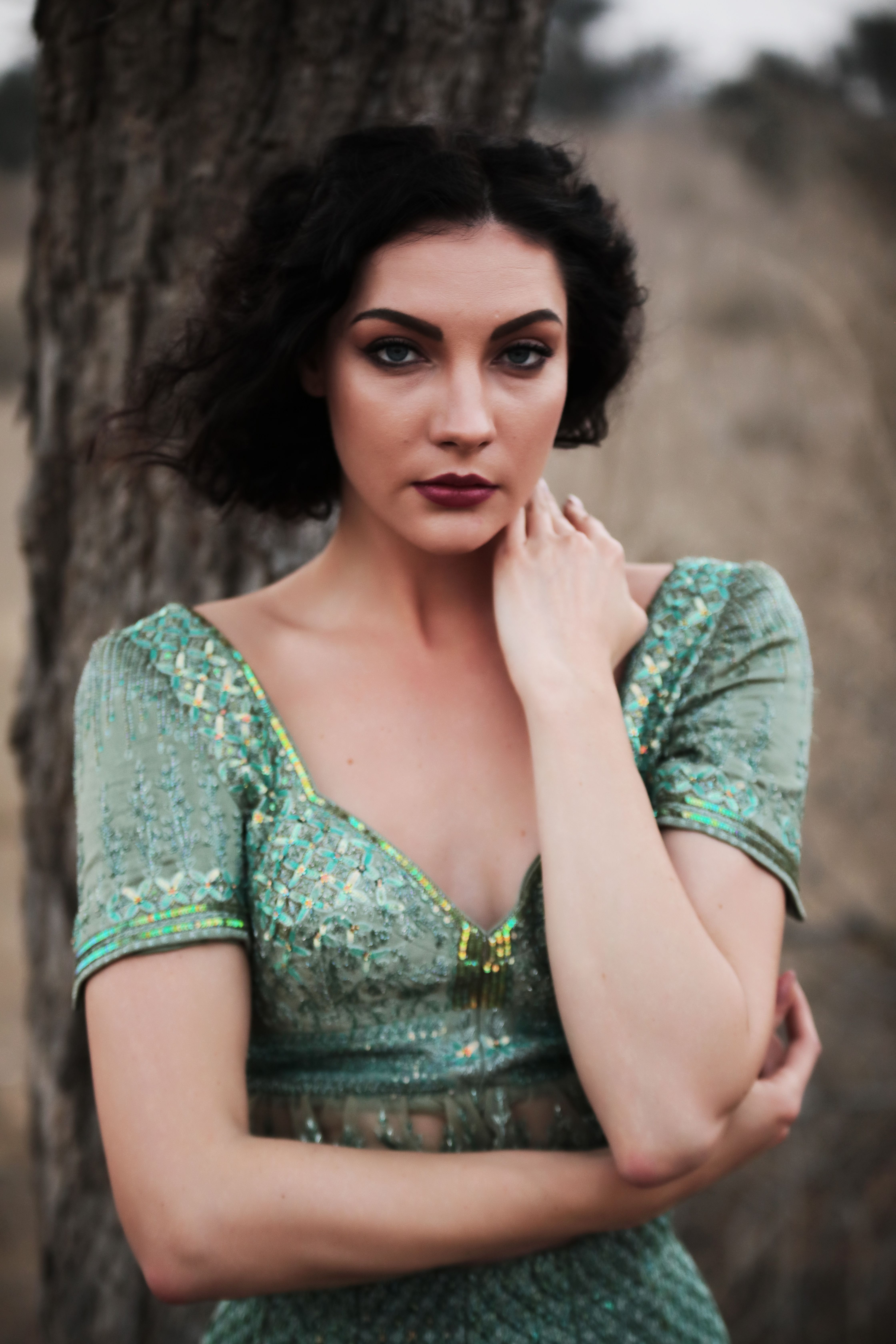 A woman in an ornate green dress poses elegantly against a tree.
