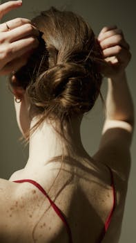 A woman adjusting her hair in a bun, casting shadows on her freckled skin, conveying elegance and simplicity.