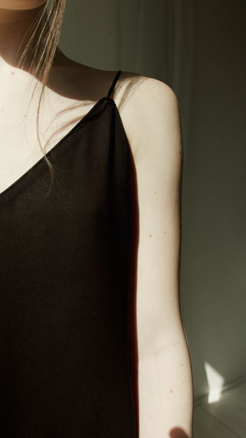 Free Close-up of a woman's shoulder in a black spaghetti strap dress with sunlight casting a soft shadow. Stock Photo