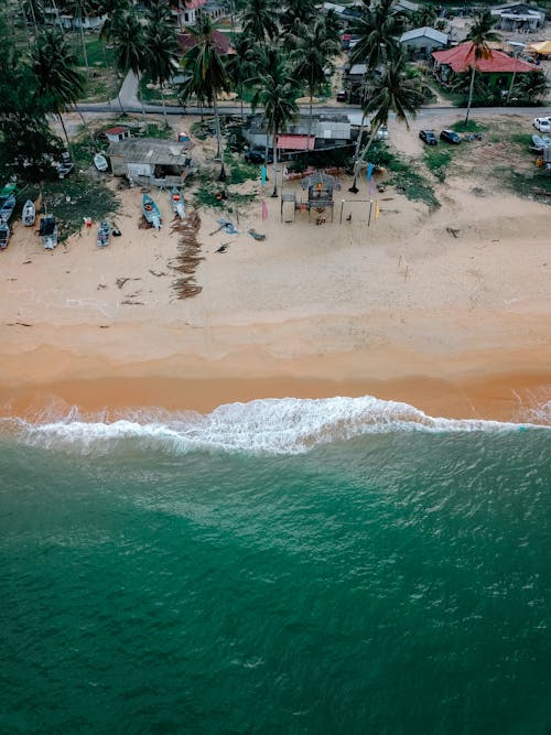 Amazing drone view of turquoise sea with white foamy waves rolling on sandy beach in tropical countryside with small houses land green palms
