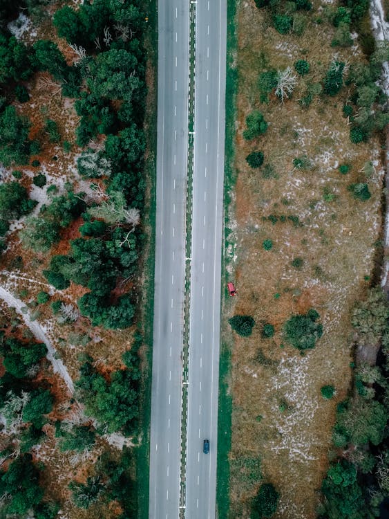 Aerial view of lonely car driving along asphalt road going through rural terrain with lush green trees and dry meadows