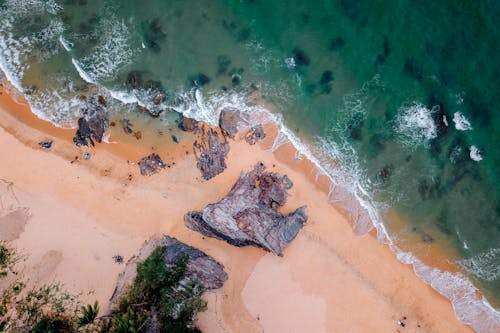 Aerial view of rocky cliffs on sandy shore of powerful foamy ocean on sunny day