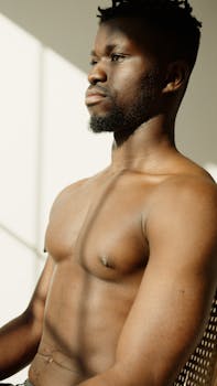 A black man sitting shirtless in natural light, highlighting muscles.