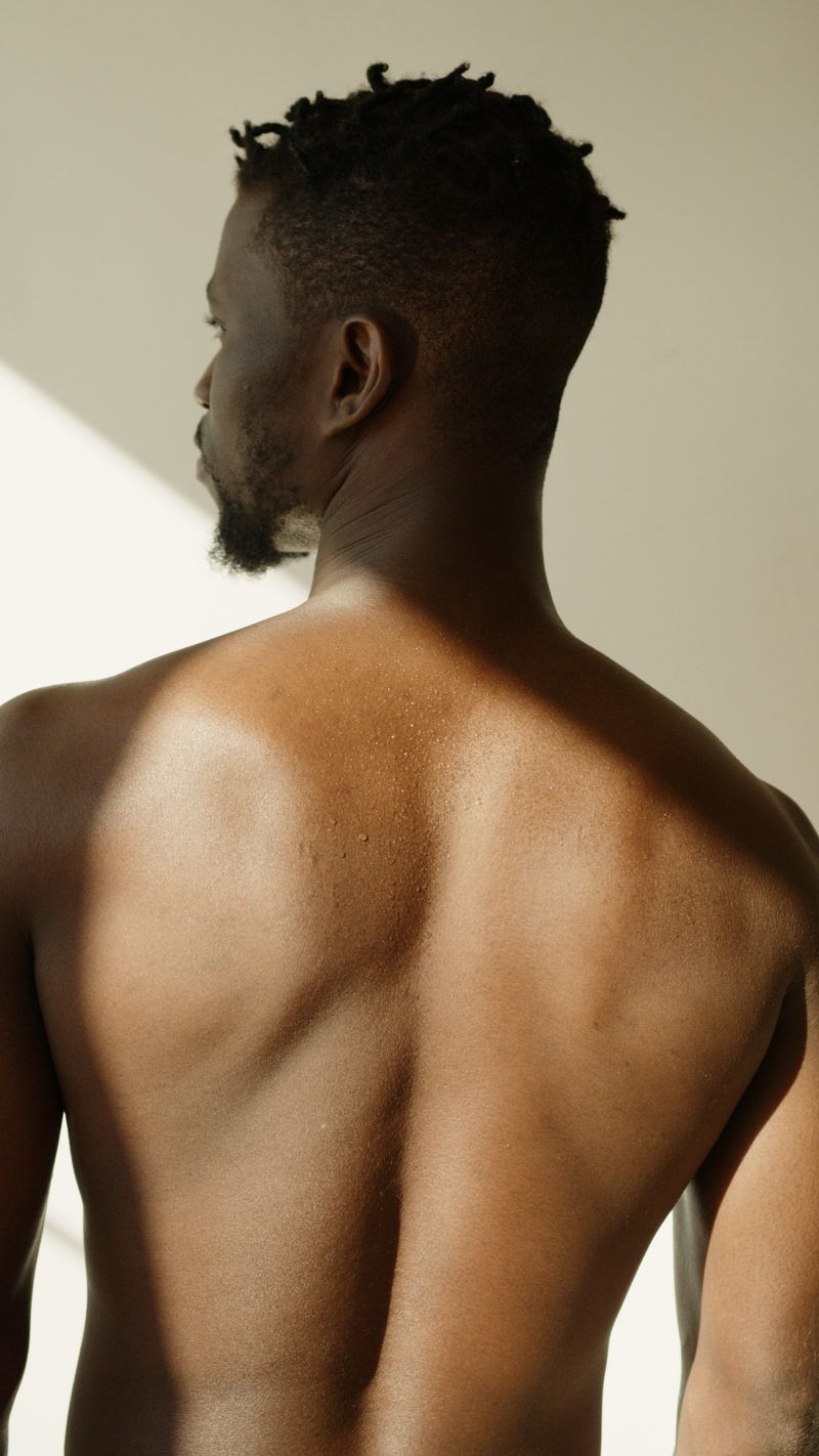 A Black man photographed from behind in warm natural light, the bare skin of his upper back and shoulders filling the frame, the area of the body where tinea versicolor most often appears.