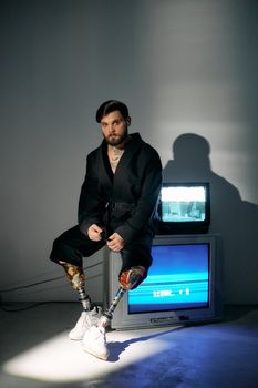A man with prosthetic legs and a beard sitting on a CRT television in a moody studio setting.