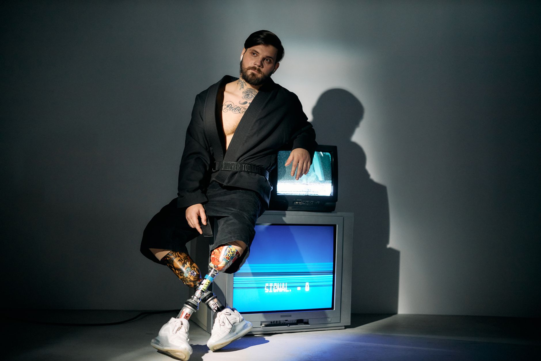 A bearded man in a bathrobe sits pensively with prosthetic legs, reflecting on technology.