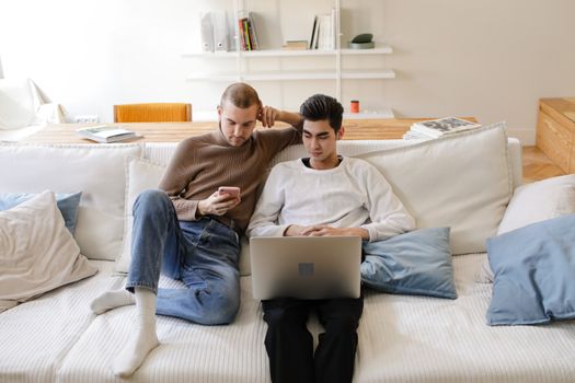 Two men relaxing on a sofa with a laptop and smartphone in a modern living room.