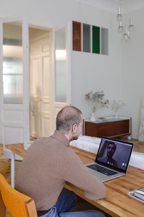 Free A man sits at a wooden table video calling a friend in a well-lit home office. Stock Photo