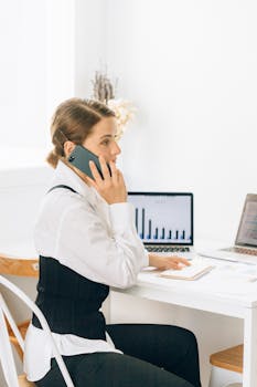 Businesswoman in corporate attire making phone call, working on laptop with graphs.