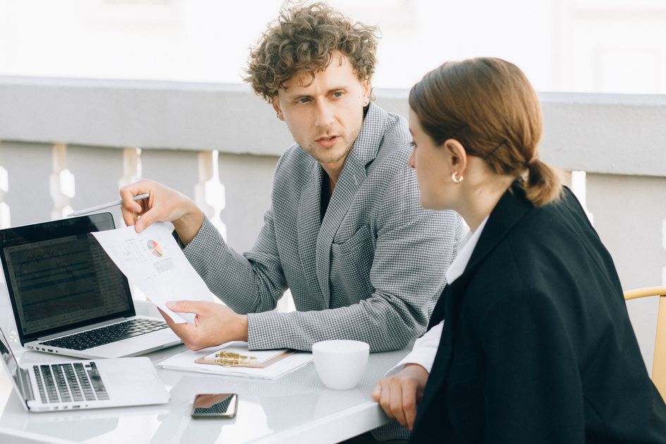Un conseiller montre un document imprimé à une cliente, assis à une table avec un ordinateur portable ouvert.