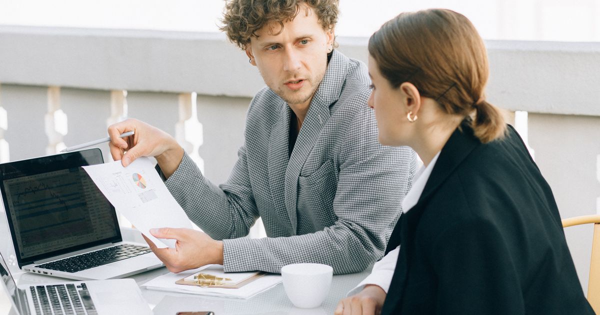 Un conseiller montre un document imprimé à une cliente, assis à une table avec un ordinateur portable ouvert.