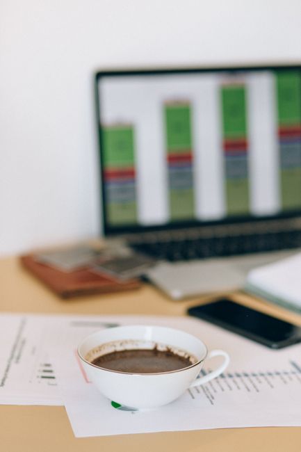 A cup of coffee sits on a desk with documents and a laptop displaying charts in the background.